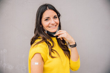 Portrait of young woman on gray wall showing medical patch after getting vaccine against...