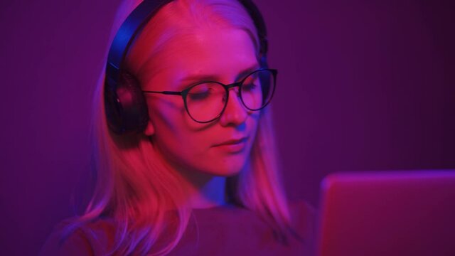 Portrait of a young female gamer at the computer club. A beautiful blonde woman in headphones and glasses sits at a computer in color light. 4K UHD