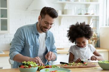 Loving young Caucasian father and small African American daughter have fun cook healthy salad together. Happy caring dad and little girl child prepare vegetarian food. Adoption, custody concept.