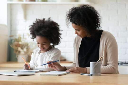 Loving young African American mother help little biracial daughter with homework assignment at home. Caring ethnic mom and small girl child study learn together. Education, homeschooling concept.