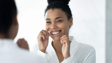 Happy Black girl in bathrobe cleaning white teeth with floss, mint thread. Young woman enjoying...