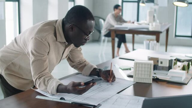 Arc shot of young Afro-American male architect leaning on office table, using smartphone and drawing floor plan on paper with pencil