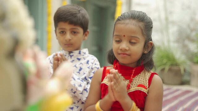 Two Kids offering prayers by clapping or Bhajan in front of god idol during Ganesha Chaturthi festival ceremony at home - concept of beliefs in Indian religious festival celebrations