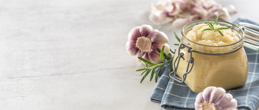 Aromatic garlic paste in a glass jar laid on rustic kitchen cloth with bulbs and peeled cloves and rosemary