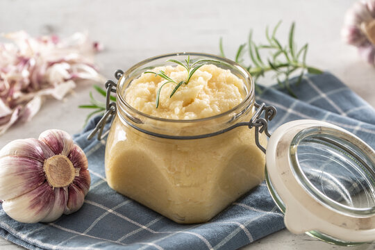 Aromatic garlic paste in a glass jar laid on rustic kitchen cloth with bulbs and peeled cloves and rosemary