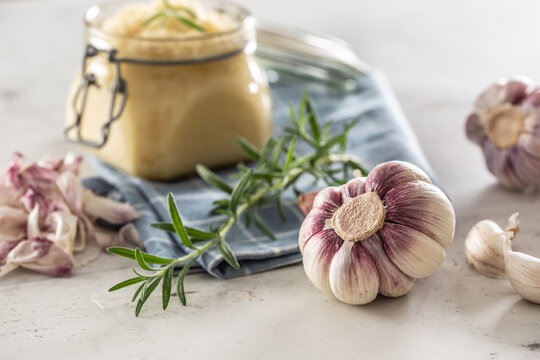 Homegrown garluc bulbs in front of a glass jar full of fresh garlic paste laid on kitchen cloth with rosemary