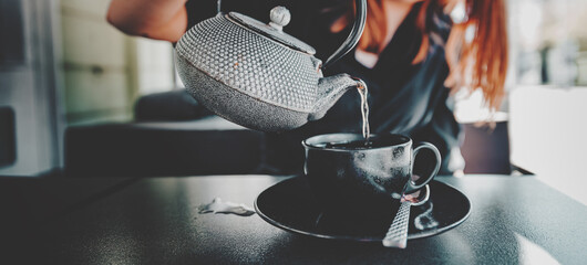 woman hand with metal teapot pour black tea in black glass cup in cafe