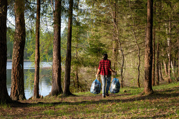 Young man with bags of trash cleaning the green forest from household rubbish, environmental...