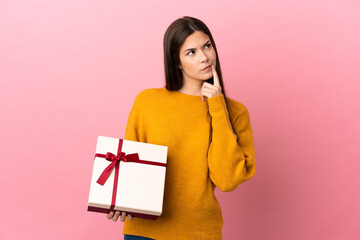 Teenager Brazilian girl holding a gift over isolated pink background having doubts while looking up