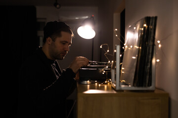 Young unshaven handsome hipster guy puts tonearm on vinyl record on turntable for sound meditation. Relaxed lounging at home with vinyl record player. Musical relaxation concept. anti-digital hobby