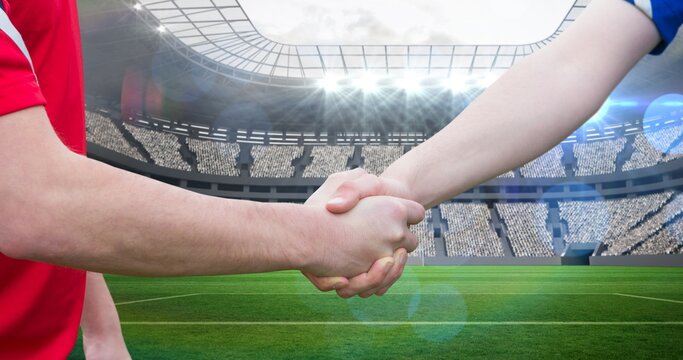 Composition of two male football players shaking hands on stadium over light blur