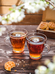 Tea party on a wooden table with branches of blossoming cherry. Black tea in glass mugs. Cherry flowers in a cup. Wooden chest with cookies in the background. Side view.