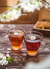 Tea party on a wooden table with branches of blossoming cherry. Black tea in glass mugs. Cherry flowers in a cup. Wooden chest with cookies in the background. Side view.