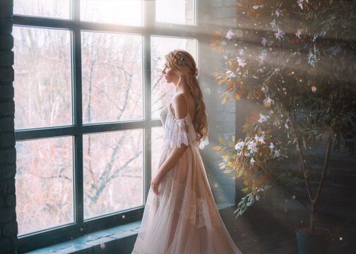 Romantic lady, blonde woman with long hair in white vintage dress stands in dark room, looks out window. Girl bride princess in wedding dress. Elegant hairstyle. Bright rays of sun concept of waiting