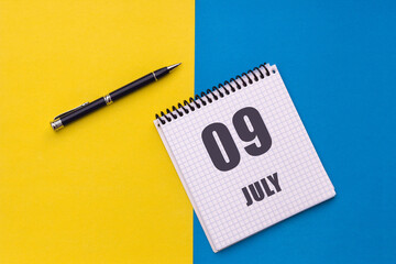 July 9th. Day 9 of month, calendar date. Notebook with a spiral and pen lies on a yellow-blue...