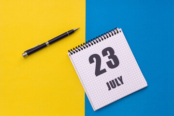 July 23th. Day 23 of month, calendar date. Notebook with a spiral and pen lies on a yellow-blue background