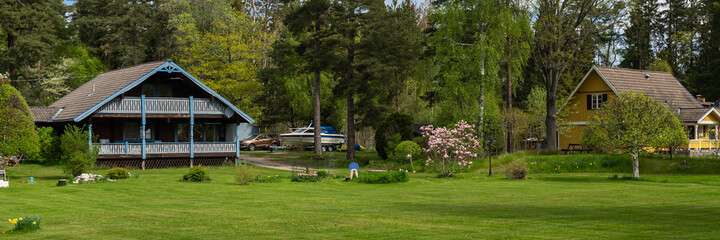 Panorama of countryside. Old two story wooden house by the road.Villa cottage made of dark wood with large terrace and balcony. Garden design with flowering trees and daffodils. Green lawn is mowed.