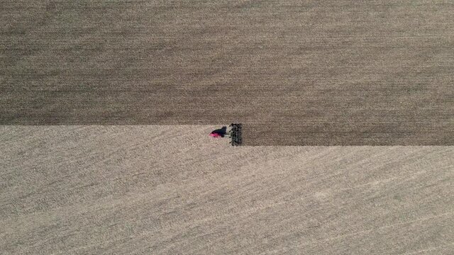 Aerial view of red tractor tilling center of farm field in the fall, sharp contrast shown on land. Driving horizontally across frame. Drone zooms in. North Dakota. Outdoors. 