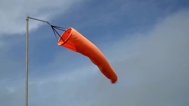 Close up of a vivid orange windsock on left side of frame blowing around in the wind. Blue sky with swirly white clouds as background. Slow motion footage of signal for transport