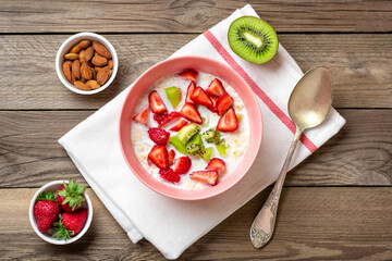 oatmeal porridge with slices of kiwi, strawberries, almonds in pink bowl, spoon, napkin with red stripes on wooden background	