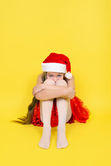 Fototapeta na wymiar Sad teen girl in red dress and Santa Claus red hat sits and hug her knees. Girl in Santa hat isolated on yellow background
