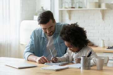 Loving young Caucasian father and African American adopted daughter write in notebook do homework assignment together. Caring dad study learn at home with biracial girl child. Adoption concept.