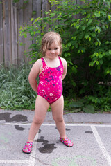 Vertical full length view of cute blond dishevelled and wet chubby toddler girl wearing a pink bathing suit pouting while standing outdoors in alley