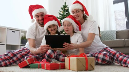 family, winter holidays and technology concept - happy mother, father and two daughters in santa hats with christmas gifts sitting on floor and having video call on tablet pc computer at home - Powered by Adobe