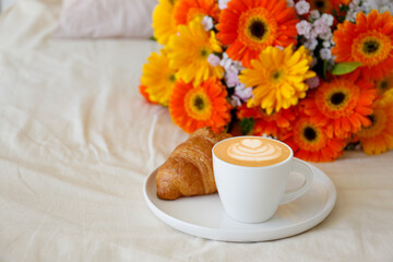 Breakfast in bed concept. White plate with cup of cappuccino and french croissant on beige sheets and colorful gerbera flowers on nightstand. Close up, top view, background, copy space for text.