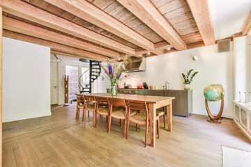 Cozy light room with wooden floor and beamed ceiling above wooden dining table and chairs in daylight