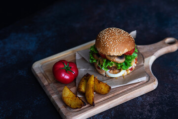Fresh craft beef burger with vegetables, mushroom, lettuce, tomatoes, cheese and sauces on a wooden rustic board on a black background. Delicious grilled home made hamburger. Fast food background.