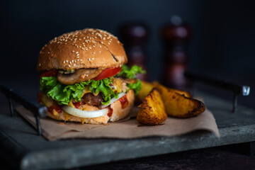 Tasty grilled homemade beef burger, tomato, cheese, cucumber and lettuce with french fries on a wooden background. Burger on the board on a dark background.
