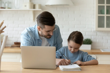 Caring young Caucasian dad and small daughter write in notebook study online on laptop at home together. Loving father and little girl child have distant lesson, prepare homework. Homeschool concept.