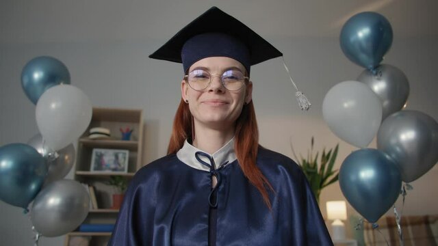 charming female graduate in academic dress is happily video chatting on laptop during online graduation ceremony while sitting at home during distance education,