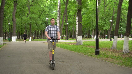 A young European man in a shirt and shorts rides an electric scooter in the park.