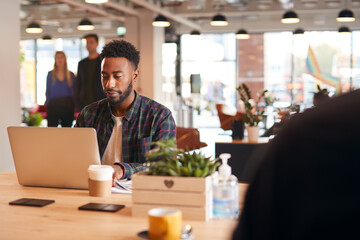 Young Businessman Sitting At Desk With Laptop In Modern Open Plan Office