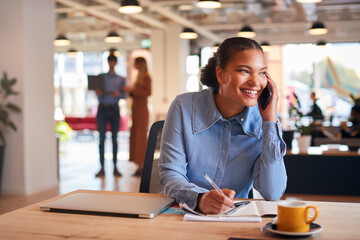 Young Businesswoman Sitting At Desk On Phone Call In Modern Open Plan Office
