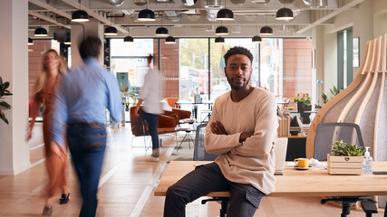 Portrait Of Businessman Sitting On Desk In Busy Multi-Cultural Office With Motion Blurred Colleagues