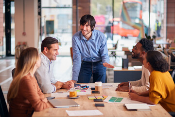 Businessman Giving Presentation To Colleagues Sitting Around Table In Modern Open Plan Office