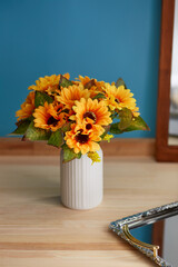 Still life photo of white ceramic vase with bunch of orange sunflowers against blue wall. Scandinavian bowl with ribbed surface is located on wooden table near silver server and mirror.  