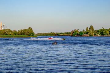 People ride PWCs and a boat on the Dnieper River in Kherson (Ukraine). Beautiful landscape with tourists frolicking in the water against the background of a green summer coastline