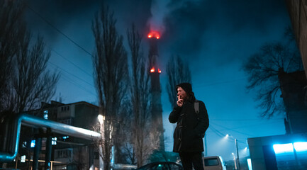 Shoot of a smoking modern man in hooded coat and lighted with neon color in the night industrial zone on background.