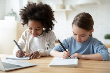 Smart small diverse girls kids write in exercise book study online on laptop together at home....