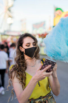 Chica joven con mascarilla por covid 19 tomando un algodón de azucar azul y tomando selfies con el movil en una zona de atracciones de una feria