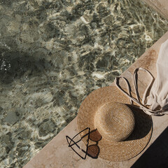 Flat lay of sunglasses and straw hat on marble swimming pool side with clear blue water with waves...