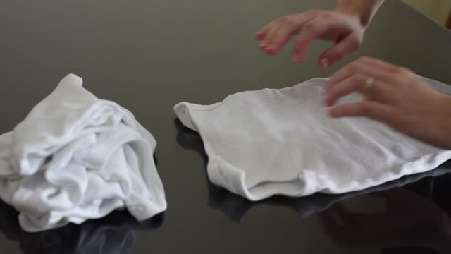 Woman's hands folding clean white baby undershirts on a dark laundry table at home. Placing in neat folded pile.