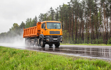 Orange dump truck with 4 axles 40 tonne truck driving on a wet road against the background of a forest, industry. Copy space for text, tonnage - obrazy, fototapety, plakaty