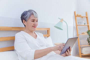 Smiling middle-aged Asia woman stay in bed browsing wireless Internet on tablet at home, happy modern senior female using pad device, elderly technology concept.