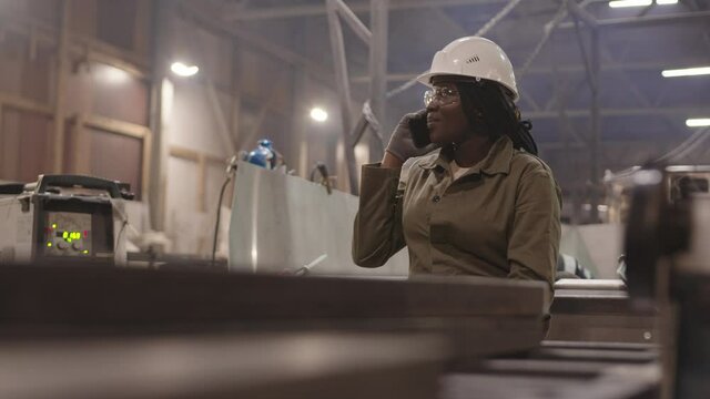 Waist-up of young African female construction worker wearing protective goggles, hard hat and gloves, sitting on edge of metallic boat, talking on mobile phone