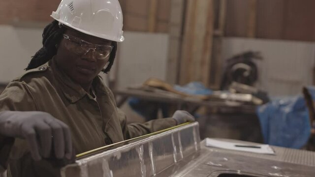 Medium close-up of female African worker wearing white hard hat and safety goggles, using tape measure on metallic piece, then writing down dimensions